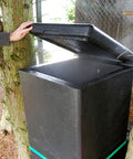 Person opening a black compost bin next to trees