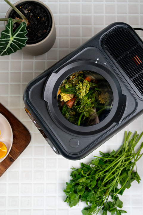 A black countertop food waste compactor filled with broccoli, cauliflower, and carrots, shown from a top-down angle on a white tiled kitchen counter with parsley and a potted plant nearby.