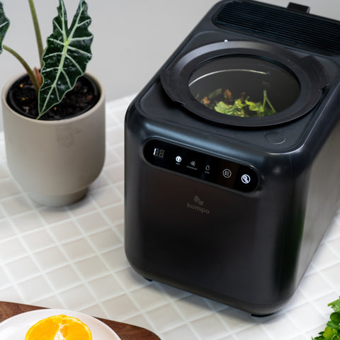 A black countertop electric composting bin with a transparent top window showing green leaves inside, placed on a white tiled surface beside a potted plant and a plate of orange slices. The front has a control panel and a visible Kompo brand mark.