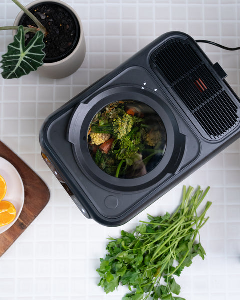 A black countertop food waste compactor filled with broccoli, cauliflower, and other vegetables, shown from above on a white tiled surface with parsley, oranges, and a potted plant nearby.