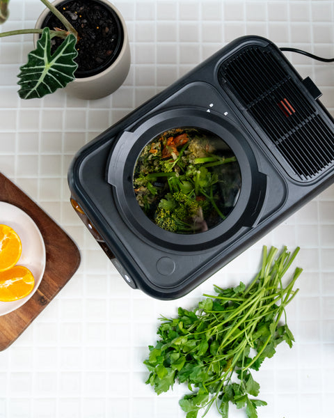 Electric composter with food on a white surface with greens and oranges.