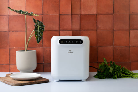 White electric food waste composter on a table with a plant and decorative items against a terracotta tiled wall.
