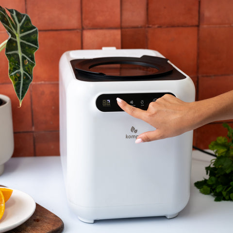 White kompo electric composter on a table with a person's hand reaching towards it, against a terracotta tiled wall.