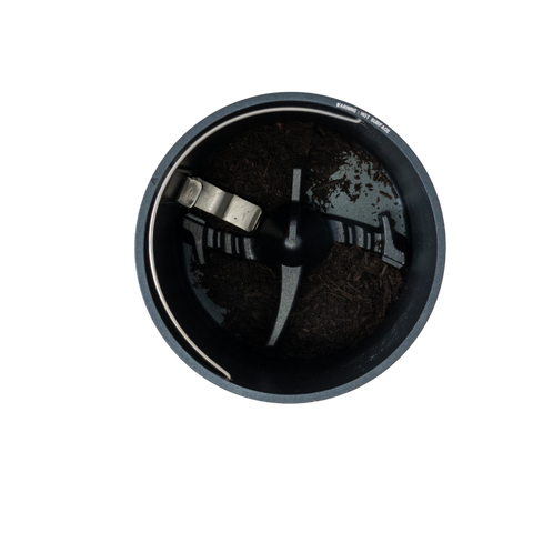 Top-down view of a circular dark gray grinder chamber with a metal blade and dark brown processed food waste inside, isolated on a white background.