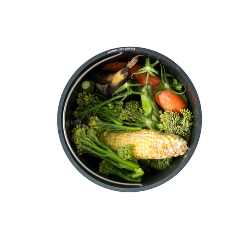 A top-down view of a round black container filled with fresh vegetables, including broccoli, carrots, and a cob of corn, isolated on a white background.